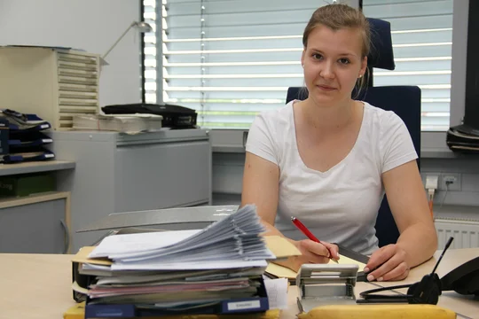 Eine Frau sitzt an einem Schreibtisch und schreibt etwas auf einem Blatt Papier
