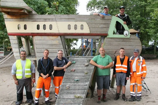 Mitarbeiter des Bauhofes stehen auf einem Spielplatz vor einem Spielgerät welches sie aufgestellt haben
